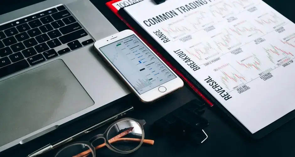 Workstation with laptop, smartphone, eyeglasses, financial charts, and pen for trading analysis.