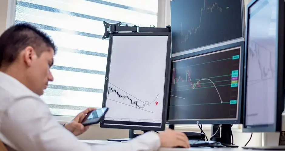 A stock trader analyzes financial data on multiple computer screens in an office setting.