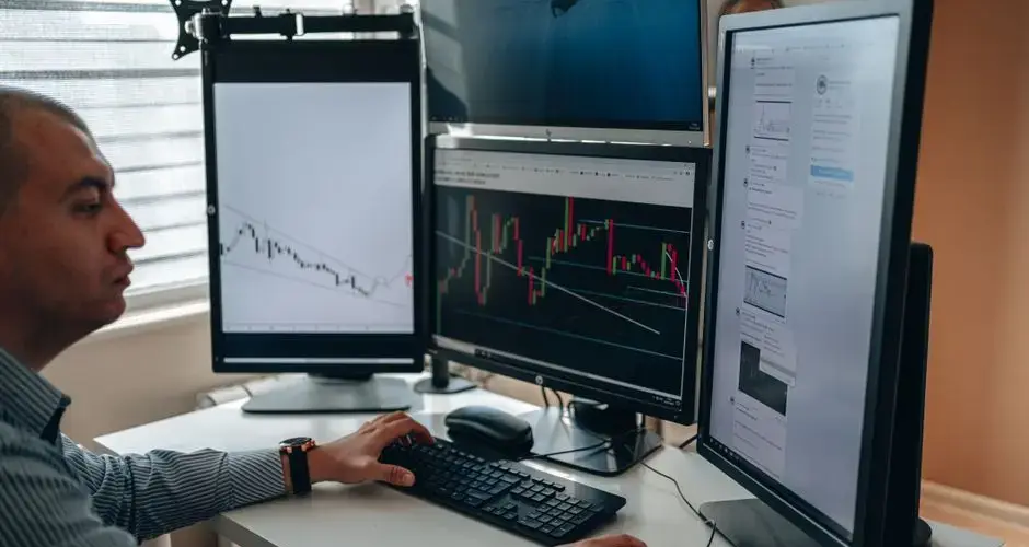 A professional in an office analyzing financial charts on multiple monitors, using advanced technology.