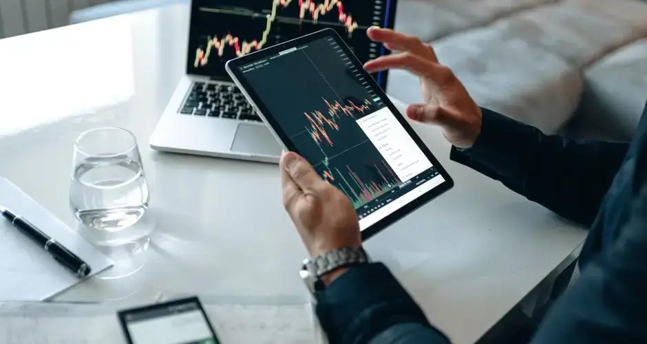 Close-up of a person using a tablet for online trading analysis with a laptop and glass of water nearby.