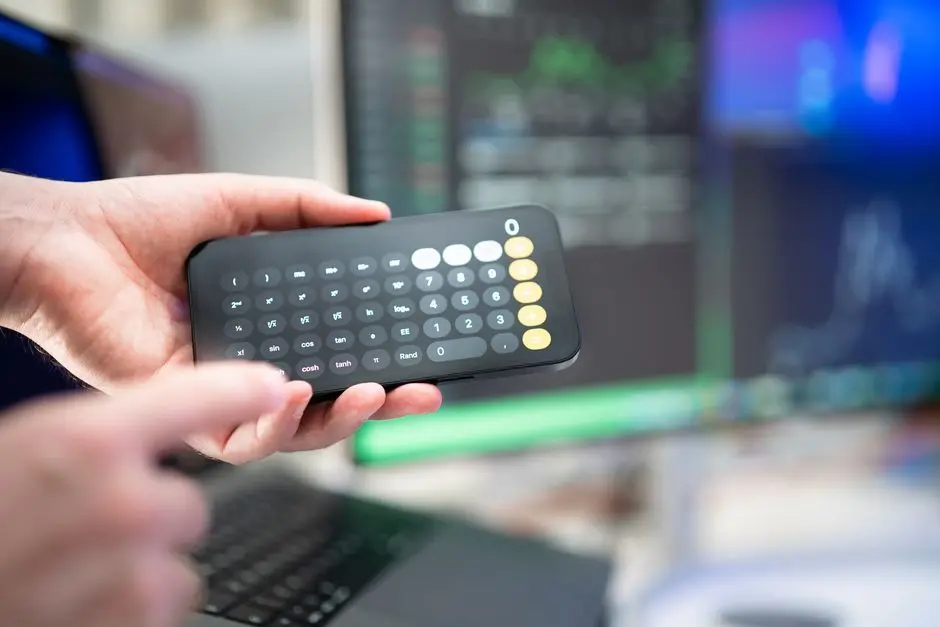 Close-up of hands using a smartphone calculator amidst computer screens in an office.