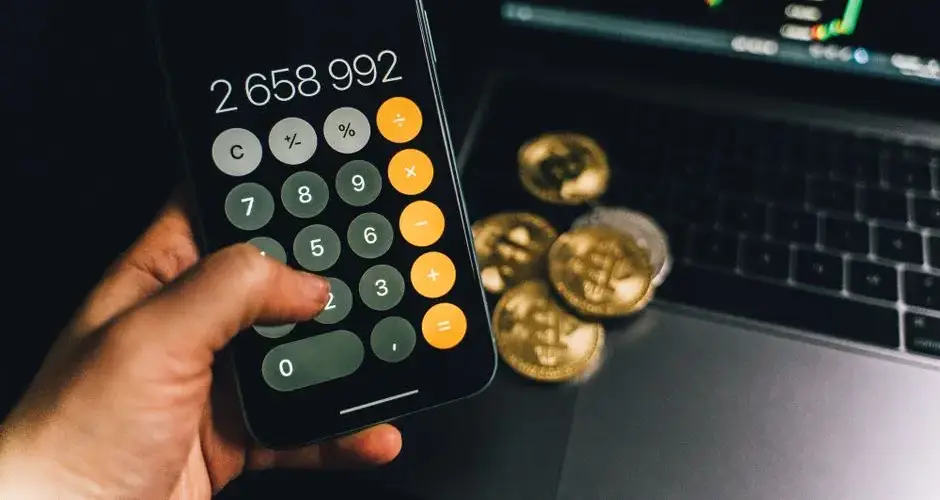 A smartphone displays a calculator app beside bitcoins and a laptop with a financial chart.