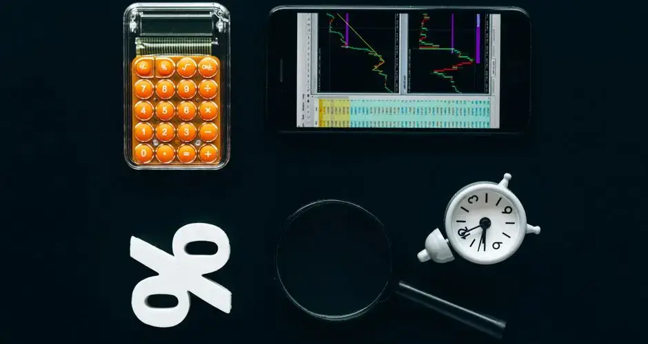 Top view of business tools including a calculator, smartphone with charts, magnifying glass, and clock on black background.