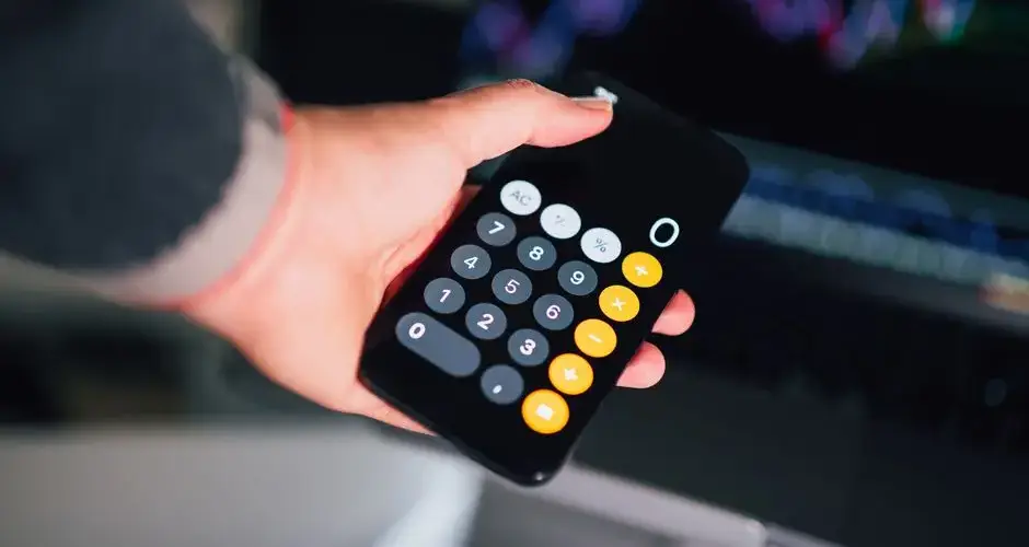 Close-up of a hand holding a smartphone calculator with financial charts on a screen in the background.