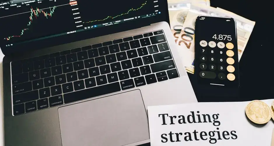 A laptop showing stock market graphs with a smartphone calculator, euro bills, coins, and trading strategies paper.