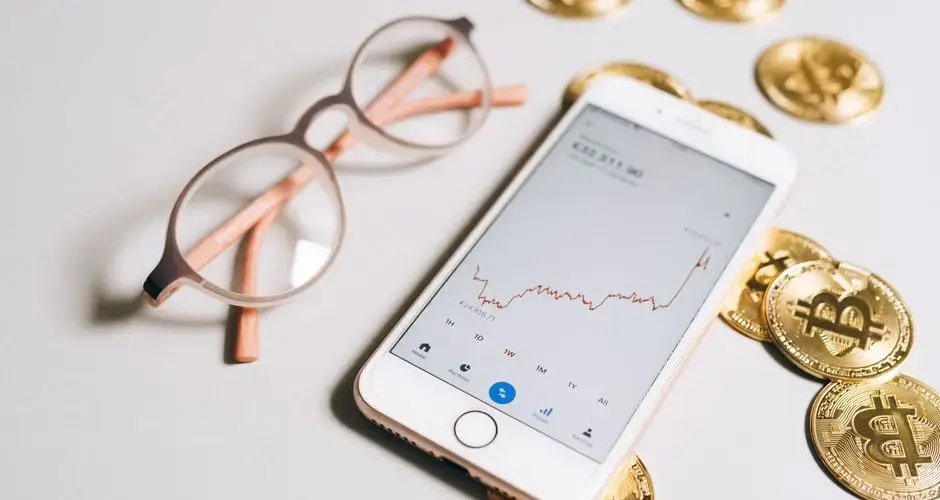 Smartphone displaying cryptocurrency graph alongside bitcoin coins and eyeglasses.