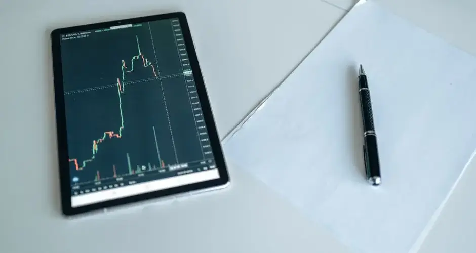 A tablet showing a stock market graph next to a pen and paper on a white surface.