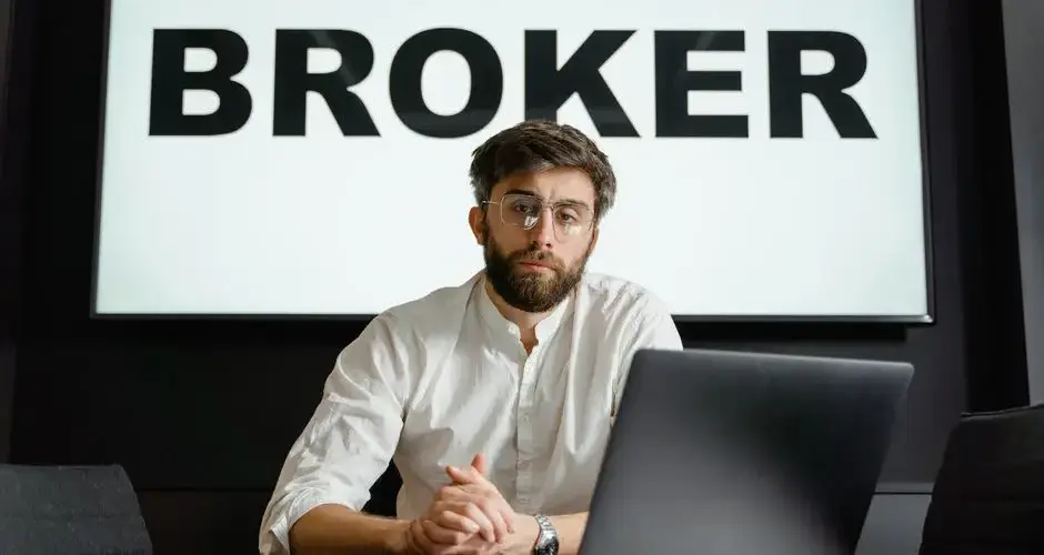 Professional bearded man in white shirt working at a laptop in a brokerage office setting.