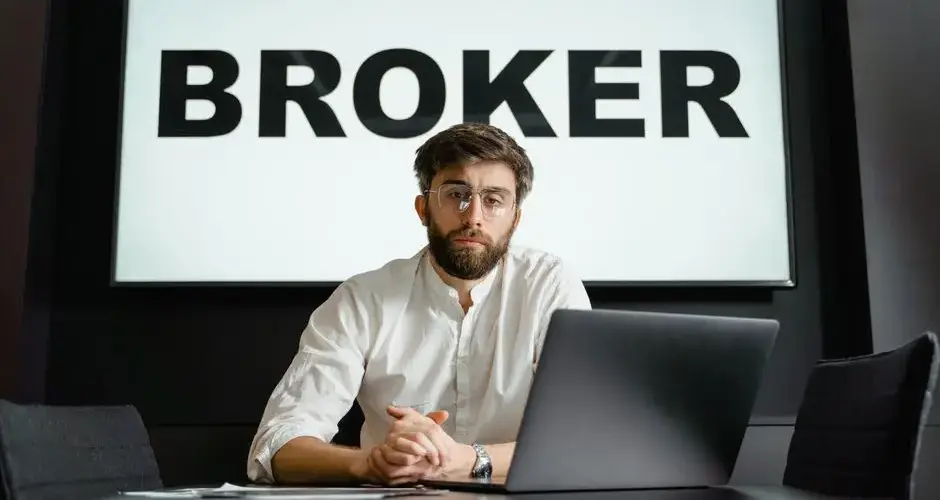 A broker with glasses sits confidently in front of a laptop in a modern conference room.