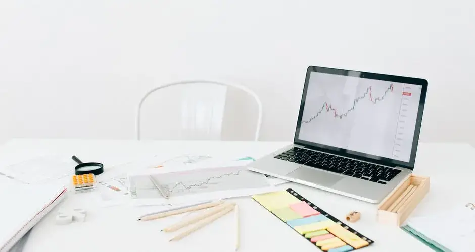 A modern workspace featuring a laptop displaying financial stock charts with documents and stationery on a white desk.