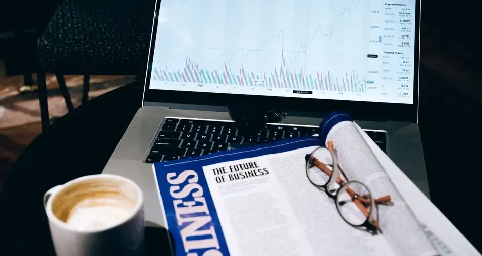 A modern business workspace featuring a laptop showing stock data next to a business magazine and coffee.