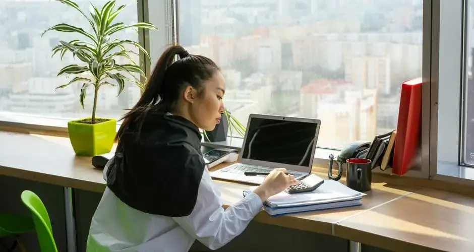 Asian woman focusing on work in a modern high-rise office setting with city view.