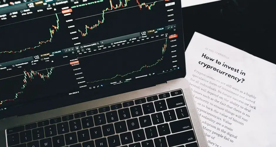 Close-up of a laptop displaying stock market graphs with a paper on investing in cryptocurrency, indoors.